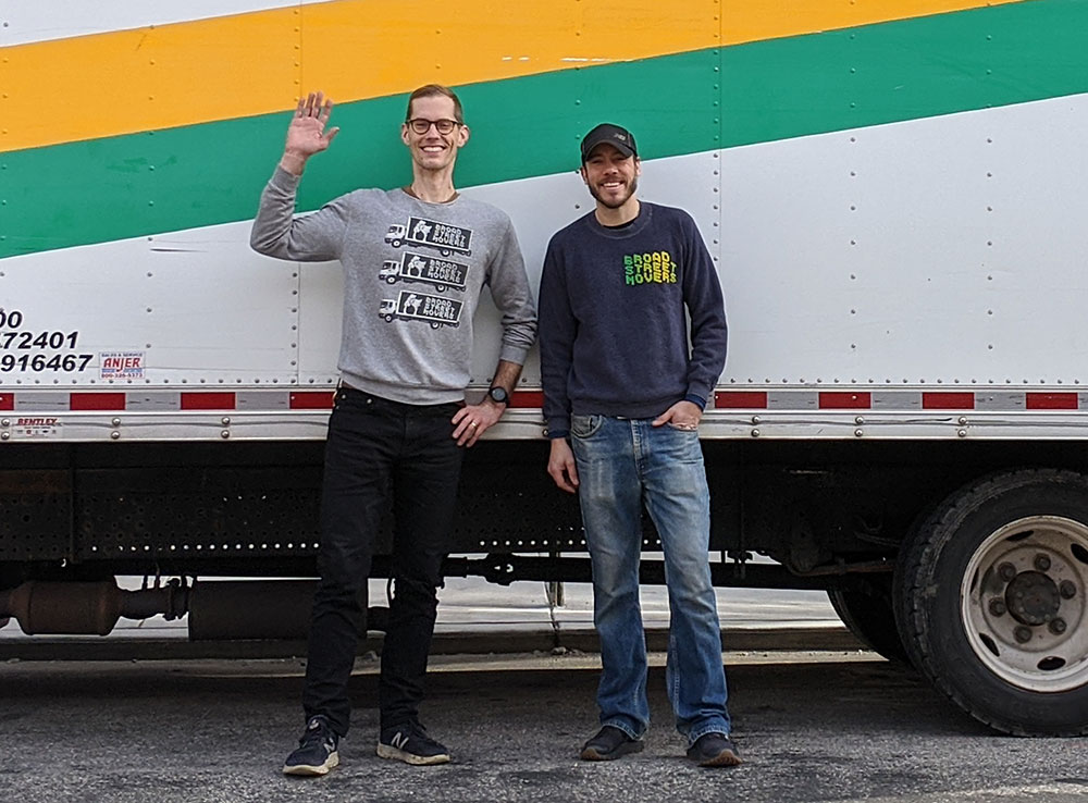Broad Street Movers team members smile cheerfully and wave hello while standing in front of a moving truck, showcasing our friendly and welcoming spirit