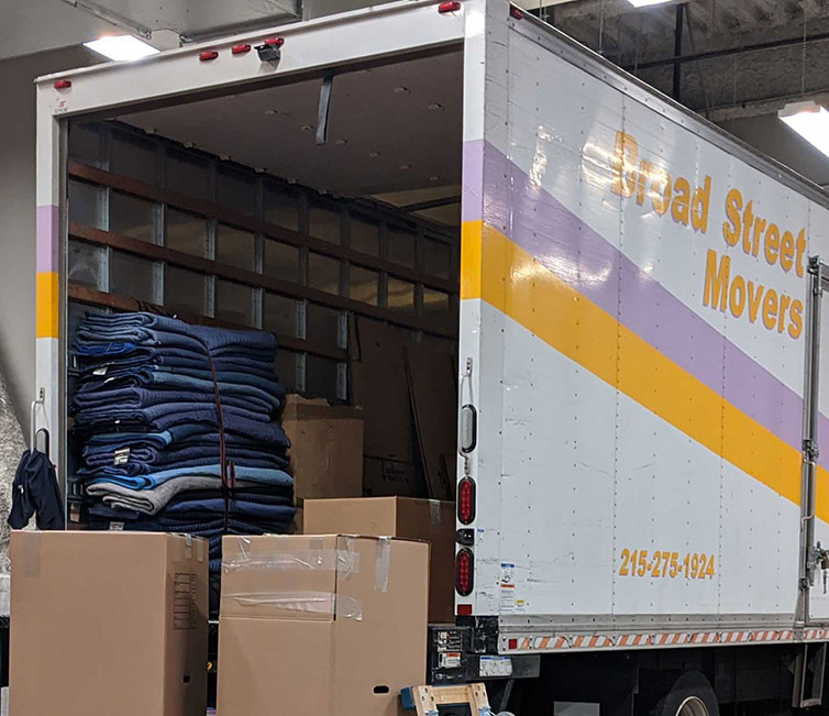 A truck from Broad Street Movers parked inside a warehouse with the back of the truck open and several tall boxes sitting on dollies waiting to be loaded inside
